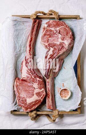 Materie non cotti black angus tomahawk bistecche su ossa servito con sale rosa sul vassoio in legno con la carta da forno sopra un panno bianco come sfondo. Vista superiore Foto Stock