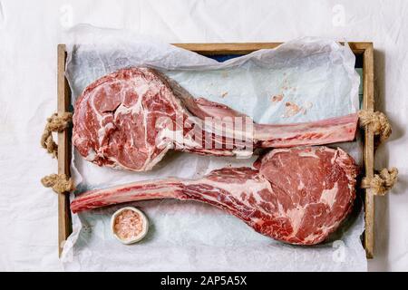 Materie non cotti black angus tomahawk bistecche su ossa servito con sale rosa sul vassoio in legno con la carta da forno sopra un panno bianco come sfondo. Vista superiore Foto Stock