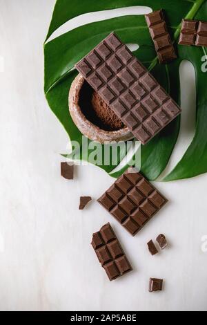 Cioccolato fondente e al latte intero bar e tritate con la polvere di cacao nel recipiente di ceramica su esotiche foglia di monstera su marmo bianco dello sfondo. Laici piana, spazio Foto Stock