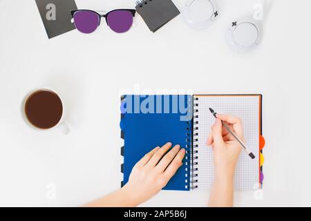Flatlay con le mani femminili che scrivono in un notebook. Spazio di lavoro minimo per l'ufficio. Sfondo bianco e tazza di caffè nero accanto. Foto Stock