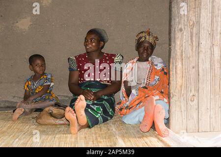 Due donne malawiche a piedi nudi e un bambino si siedono su una canna tappetino al piano della casa del villaggio con abiti tradizionali Foto Stock