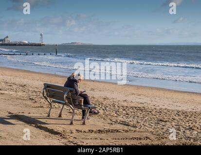 Bournemouth Beach con una persona seduta da sola sul lungomare mentre si utilizza un telefono cellulare in inverno. Foto Stock