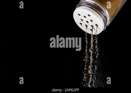 Il pepe nero macinato cade dallo shaker del pepe su uno sfondo nero isolato. Il processo di stagionatura si interrompe in tempo. Agitatore con sale di vetro con st. Bianco Foto Stock