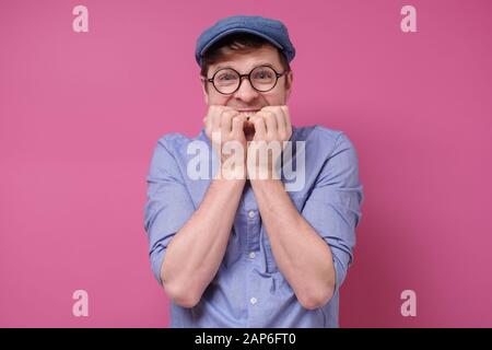 Giovane uomo divertente in occhiali e cappello mordendo le unghie sulla parete rosa. Studio Foto Stock