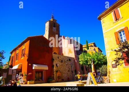 Roussillon En Provence, Francia - 2 Ottobre. 2019: Vista oltre la casa gialla mediterranea luminosa sulla torre della chiesa ocra contro il cielo blu Foto Stock