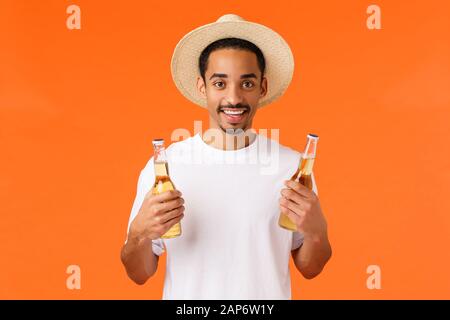 Come bere fuori. Bello rilassato e felice uomo afroamericano sorridente in cappello, maglietta bianca, tenendo due bottiglie di birra, godendo fine settimana Foto Stock