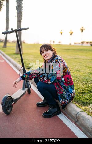 Allegra donna sorridente sta seduto vicino al suo scooter elettrico in un parco cittadino Foto Stock