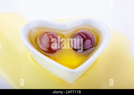 ciotola a forma di cuore su sfondo bianco con due olive scure in olio d'oliva su fondo olio d'oliva Foto Stock