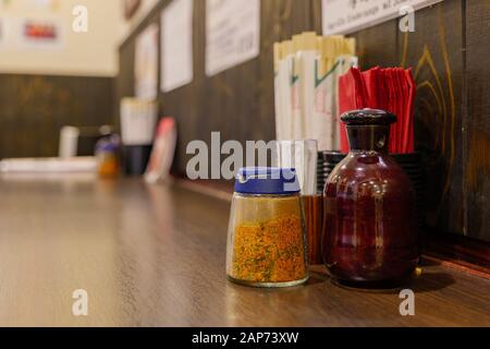 Bottiglie di stagione, con scaglie di peperoncino rosso giapponese e bottiglia di salsa di soia, tovaglioli rossi, bastoncini e stuzzicadenti su banco di legno del ristorante giapponese. Foto Stock