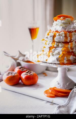 Un multistrato Pavlova torta servito con panna montata aromatizzata con terreno fresco cardamomo, purea di cachi, kaki fette. Foto Stock