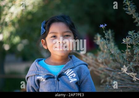 Bambina sicura che indossa un maglione, in piedi di fronte agli alberi e piante verdi con un sorriso sincero sul suo viso. Foto Stock