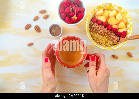 Vista dall'alto, mani femminili che tengono una tazza di caffè, un cucchiaio con farinata d'avena, con noci di miele, lamponi, su un tavolo di legno giallo Buongiorno - Foto Stock