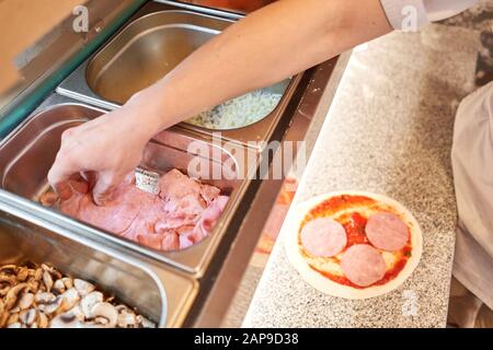 Cucinare la pizza. Presenta il ripieno di formaggio e prosciutto, gli ingredienti sulla preforma di pasta. Mano di primo piano di panettiere dello chef in uniforme grembiule bianco cucinare a Foto Stock