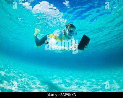 Snorkeling uomo d'affari in camicia e cravatta e pinne corrispondenti utilizzando un computer tablet mentre nuotano sotto l'acqua in mare tropicale turchese Foto Stock