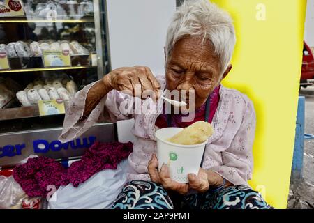 Antipolo City, Filippine - 18 gennaio 2020: Vecchia donna filippina con pelle rugosa mangiare una ciotola di porridge mentre si siede su un marciapiede. Foto Stock