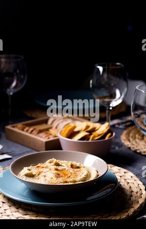 Gustoso hummus ceci salsa alimentare su un piatto su un tavolo da pranzo con tessuti blu e grigi su sfondo nero Foto Stock