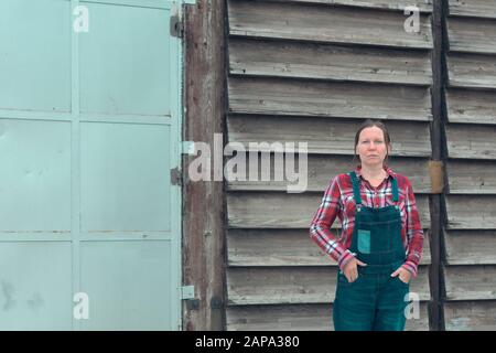 Ritratto di contadino femminile di fronte al capannone. Donna vestita con camicia plid e jeans jeans jeans tute come lavoratore fattoria con le mani in tasche Foto Stock