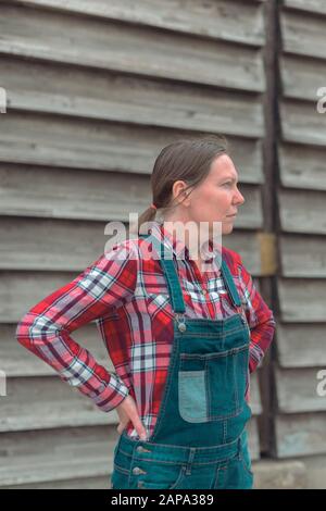 Ritratto di contadino femminile di fronte al capannone. Donna vestita con camicia plid e jeans jeans jeans tute come lavoratore fattoria con le mani sui fianchi Foto Stock