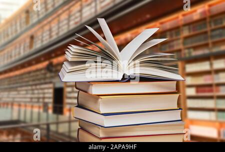 Pila di libri con un libro aperto davanti agli scaffali di una grande biblioteca Foto Stock