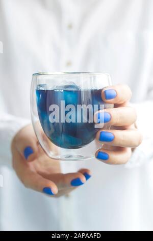 Matcha blu bevanda fredda con ghiaccio in un bicchiere di vetro nelle mani di una donna con manicure blu. Colore dell'anno 2020 trend Foto Stock