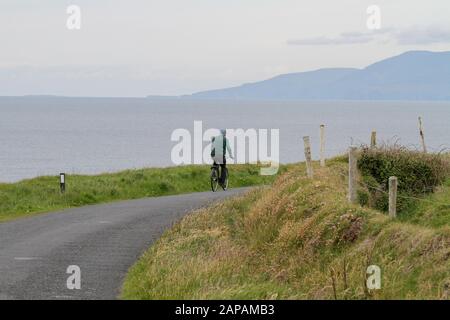 Un uomo anziano in bicicletta in Irlanda in bicicletta lungo il mare su una strada costiera rurale in una giornata grigia a Mullaghmore Head nella contea di Sligo. Foto Stock