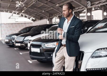 Uomo d'affari giovane e fiducioso che si trova vicino all'auto nel parcheggio Foto Stock