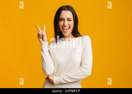 Giocosa ragazza bruna che mostra segno di pace e che si inocchiano su sfondo giallo Foto Stock