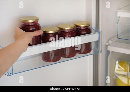 Vasetti in vetro con marmellata di lamponi in frigorifero. Mano femminile con un vasetto di marmellata rossa fatta in casa. Cibi vegetariani sani fermentati. Foto Stock