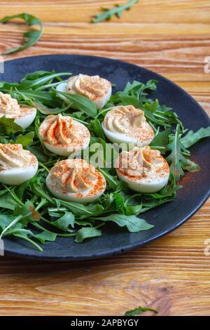 Uova divorate con paprika su insalata di rucola fresca in piatto rurale nero su tavola di legno, sano antipasto vegetariano o spuntino vicino Foto Stock