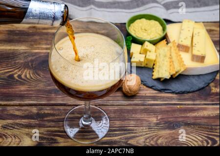 Versare la birra rossa dell'abbazia belga in vetro e piatto di pietra con formaggi prodotti da monaci di latte vaccino con fenugreek e semi di senape vicino Foto Stock