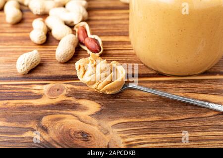 Burro di arachidi nel cucchiaio vicino alla pasta cremosa di arachidi in vaso di vetro aperto. Arachidi nella buccia sparse sul tavolo di legno marrone con spazio copia per cucinare Foto Stock