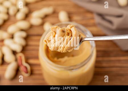 Burro di arachidi nel cucchiaio vicino alla pasta cremosa di arachidi in vaso di vetro aperto. Arachidi nella buccia sparse sul tavolo di legno marrone con spazio copia per cucinare Foto Stock