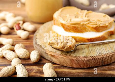 Burro di arachide nel cucchiaio vicino pasta cremosa di arachide in vaso di vetro aperto, fetta di pane di burro di arachide, pane tostato. Arachidi nella buccia sparse sul marrone Foto Stock