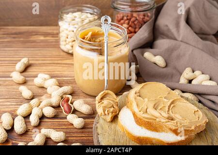 Burro di arachide nel cucchiaio vicino pasta cremosa di arachide in vaso di vetro aperto, fetta di pane di burro di arachide, pane tostato. Arachidi nella buccia sparse sul marrone Foto Stock