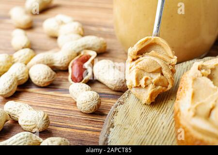 Burro di arachide nel cucchiaio vicino pasta cremosa di arachide in vaso di vetro aperto, fetta di pane di burro di arachide, pane tostato. Arachidi nella buccia sparse sul marrone Foto Stock