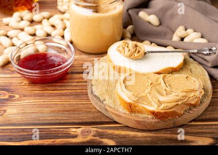 Burro di arachide nel cucchiaio vicino pasta cremosa di arachide in vaso di vetro aperto, fetta di pane di burro di arachide, pane tostato, marmellata. Arachidi in buccia sparse su legno marrone Foto Stock