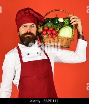 Cucinare con il viso stretto in uniforme borgogna tiene le verdure nel cesto di vimini. Lo chef tiene cavolo, ravanello, broccoli con lattuga e aglio. Concetto di cucina sana. Uomo con barba su sfondo rosso Foto Stock