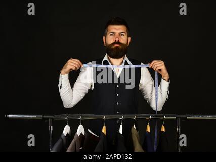 Bell'uomo con regolabarba misura il nastro vicino al collo nel negozio di abbigliamento. Sarto o venditore accanto a rack con giacche classiche. Personalizzazione e stile individuale Foto Stock