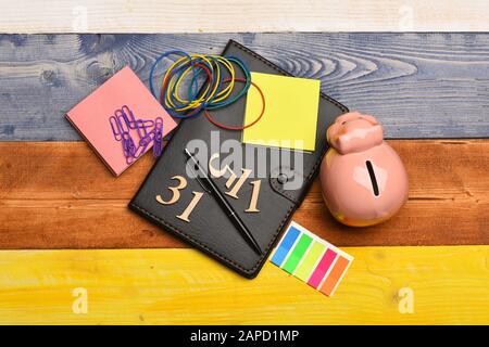 Banca piggy e cancelleria su sfondo colorato di legno. Concetto bancario e di profitto. Moneybox con la faccia di maiale per organizzatore, note adesive, segnalibri, penna, elastici e numeri. Composizione finanziaria domestica Foto Stock