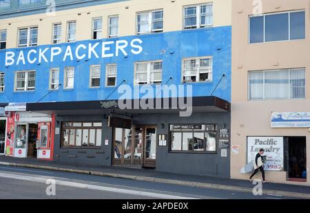 Un surfista passeggia davanti a un Bondi Backpackers Hotel, negozi fronti e appartamenti sulla Campbell Parade Foto Stock