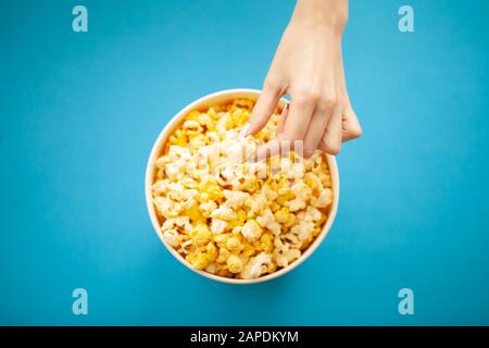 Cibo. Donna Mano che Prende Popcorn dal secchio. Benna Per Popcorn Su Sfondo Blu. Cinema Foto Stock