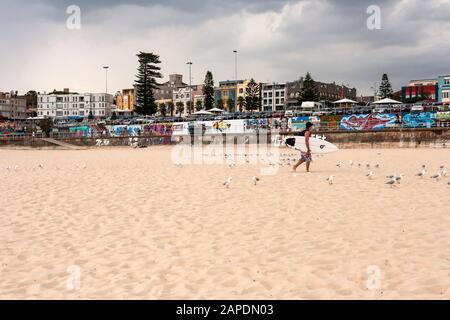 Un surfista trasporta la sua tavola da surf attraverso la famosa spiaggia di Bondi in una giornata torbida a metà estate. Foto Stock