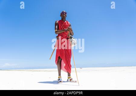 Zanzibar, Tanzania - 28 ottobre 2019 : masai africano vestito con abiti tradizionali in piedi vicino all'oceano sulla spiaggia di sabbia, Zanzibar, Tanzania Foto Stock