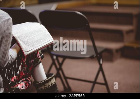 Primo piano di una persona seduta su una sedia e che legge una bibbia in una chiesa sotto le luci Foto Stock