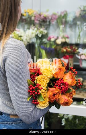 La ragazza tiene un bel bouquet di fiori d'arancio e rosso, rose, accanto al bancone dei fiori. Bella donna sceglie un bouquet in un negozio fiorista. Foto Stock