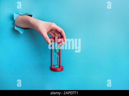 La mano femmina tiene una clessidra attraverso un foro in carta su uno sfondo blu. Concetto Di Perdita Di Tempo Foto Stock