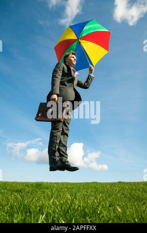 Buon uomo d'affari con ombrello colorato e valigetta galleggiante all'aperto sopra ampio prato verde Foto Stock
