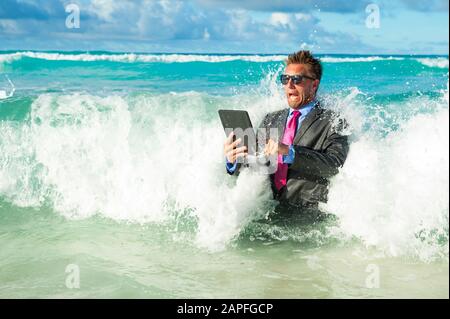 Un uomo d'affari scioccato che cerca di usare il suo computer tablet in onde che si infrangono sulla riva di una spiaggia tropicale Foto Stock