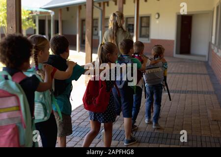 Insegnante femminile con un gruppo di scolaresche che camminano in un corridoio esterno in una scuola elementare Foto Stock