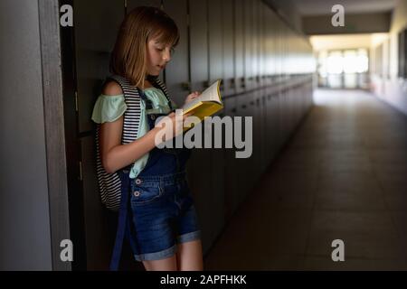 Scolaretta appoggiata contro un muro in un corridoio esterno che legge un libro alla scuola elementare Foto Stock